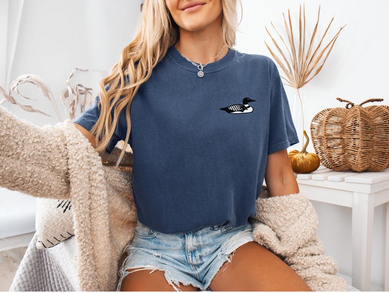 May include: A woman wearing a dark blue t-shirt with a black and white loon graphic. The shirt has short sleeves and a relaxed fit. The woman is wearing denim shorts and a cream-coloured cardigan.