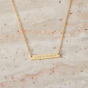 May include: A gold-colored bar necklace with a delicate chain. The rectangular pendant is engraved with the coordinates "43.3821° N, 95.1588° W". The necklace is displayed on a textured, light-colored surface.