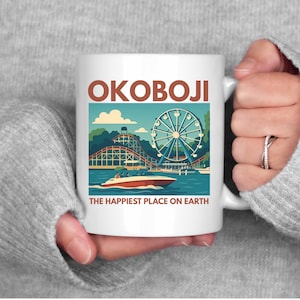 May include: White ceramic mug featuring a colorful illustration of a Ferris wheel, roller coaster, and boat on a lake. The mug has the word "OKOBOJI" at the top and the phrase "THE HAPPIEST PLACE ON EARTH" at the bottom.