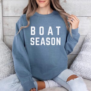 May include: A blue sweatshirt with the words "BOAT SEASON" in white block letters. The sweatshirt is a solid color and has long sleeves. The person is wearing light blue jeans.