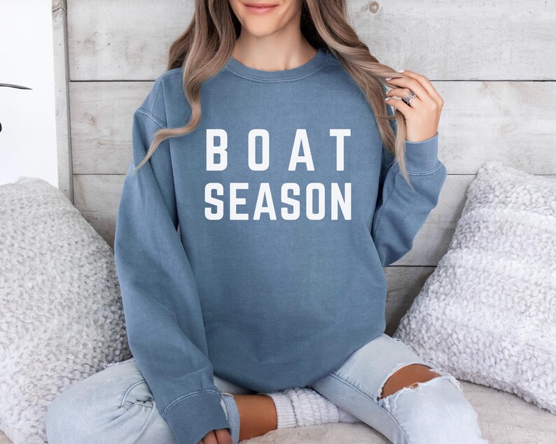 May include: A blue sweatshirt with the words "BOAT SEASON" in white block letters. The sweatshirt is a solid color and has long sleeves. The person is wearing light blue jeans.