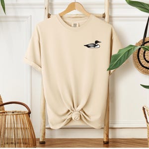 May include: A beige t-shirt with a black loon graphic on the left chest.