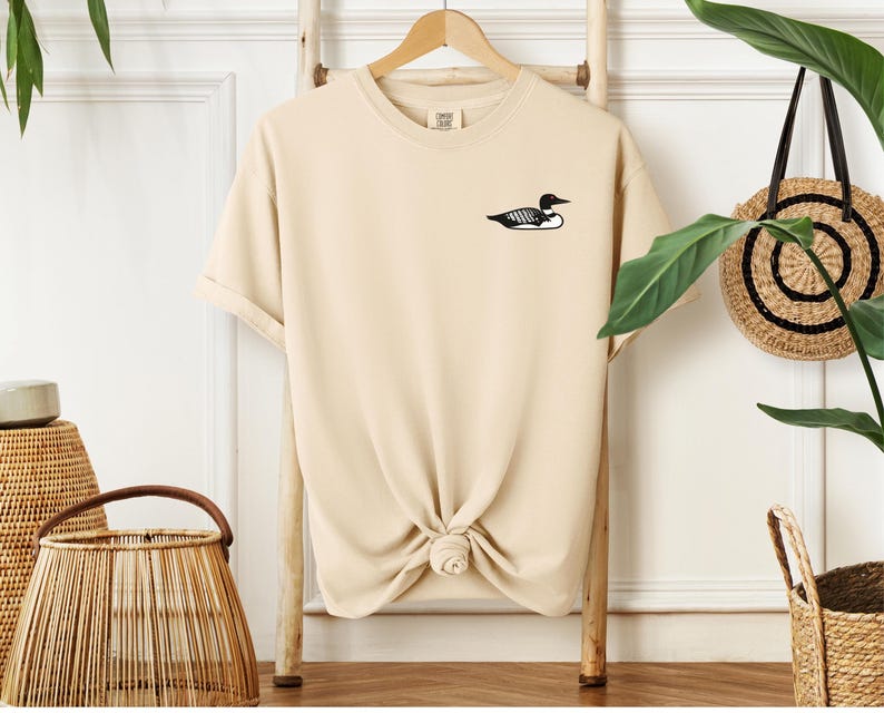 May include: A beige t-shirt with a black loon graphic on the left chest.