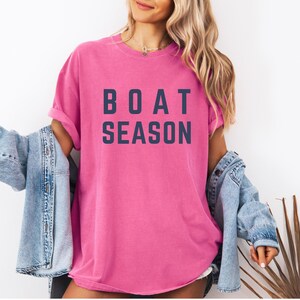 May include: A bright pink t-shirt with the words "BOAT SEASON" printed in navy blue block letters. The shirt is worn with a light blue denim jacket.
