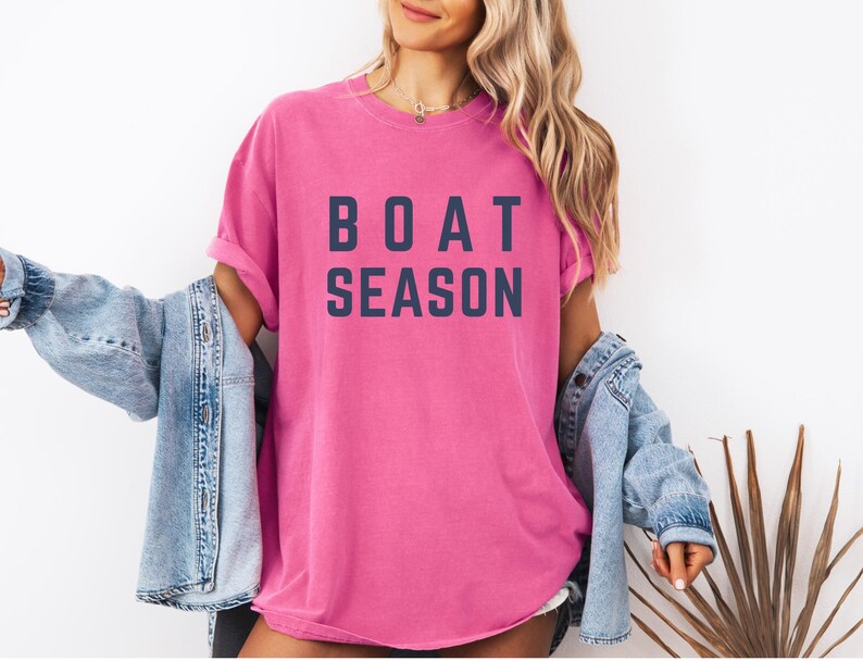 May include: A bright pink t-shirt with the words "BOAT SEASON" printed in navy blue block letters. The shirt is worn with a light blue denim jacket.