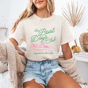 Pu&ograve; includere: T-shirt color crema con la scritta "The Boat Days Club" in verde e una grafica di una barca rosa. La frase "happiest on the water" &egrave; sotto la grafica. La maglietta &egrave; abbinata a pantaloncini di jeans.