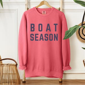 May include: A coral-colored sweatshirt with the words "BOAT SEASON" printed in navy blue block letters. The long-sleeved top is hanging on a wooden rack. The sweatshirt has a relaxed fit and rolled cuffs.