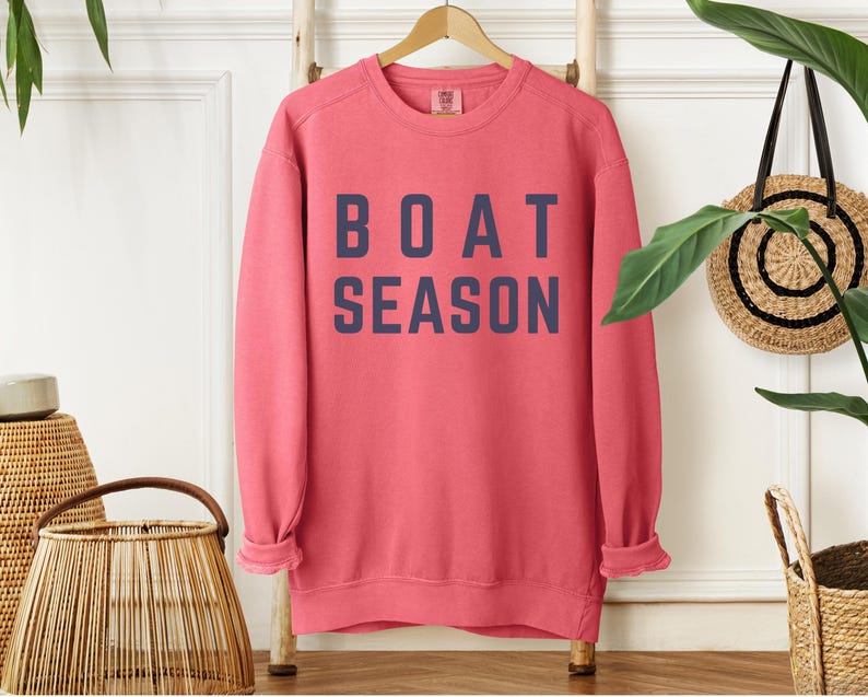 May include: A coral-colored sweatshirt with the words "BOAT SEASON" printed in navy blue block letters. The long-sleeved top is hanging on a wooden rack. The sweatshirt has a relaxed fit and rolled cuffs.