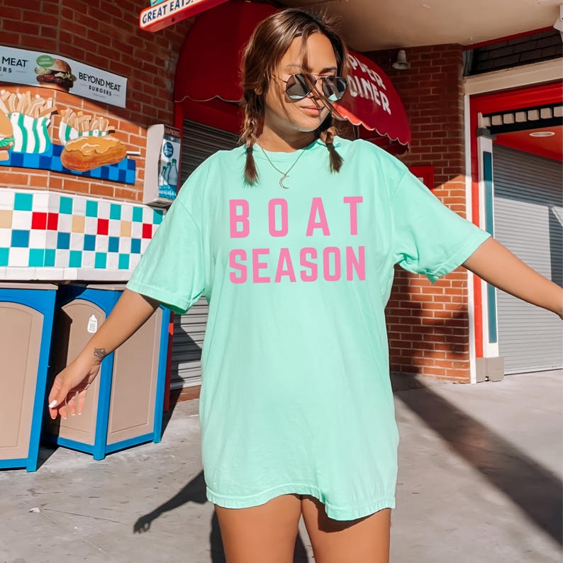 May include: A light green t-shirt with the words "BOAT SEASON" printed in pink. The shirt is oversized and appears to be made of a soft material. The person wearing the shirt is wearing sunglasses and has their arms outstretched.