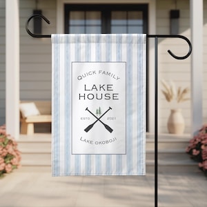 Puede incluir: Una bandera de jardín con un fondo a rayas azules y blancas. La bandera presenta un recuadro blanco con las palabras "QUICK FAMILY LAKE HOUSE" y "LAKE OKOBOJI" en texto negro. También incluye remos cruzados y un pequeño gráfico de árbol.
