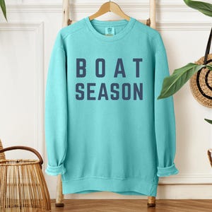 May include: A turquoise sweatshirt with the words "BOAT SEASON" printed in navy blue block letters. The sweatshirt is hanging on a wooden hanger against a white wall. A woven basket and a decorative wall hanging are also visible.