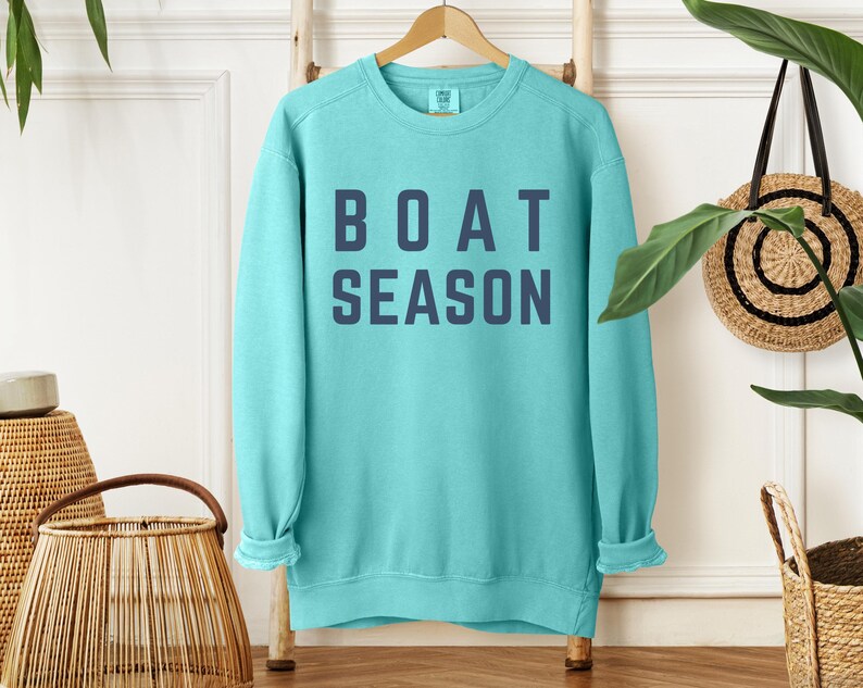 May include: A turquoise sweatshirt with the words "BOAT SEASON" printed in navy blue block letters. The sweatshirt is hanging on a wooden hanger against a white wall. A woven basket and a decorative wall hanging are also visible.