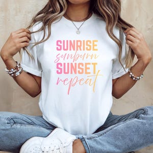 May include: White t-shirt with a graphic design that reads "Sunrise, Sunburn, Sunset, Repeat" in a gradient of pink, orange, and yellow.