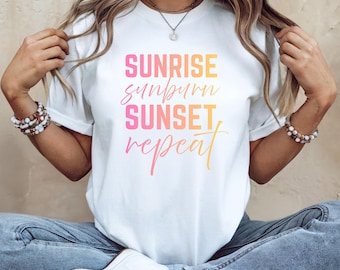 May include: White t-shirt with a graphic design that reads "Sunrise, Sunburn, Sunset, Repeat" in a gradient of pink, orange, and yellow.