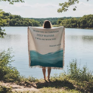 K&ouml;nnte beinhalten: Eine cremefarbene, gewebte Wohndecke mit einem blauen Aquarell-Wellenmuster am unteren Rand und dem in Dunkelblau gedruckten Text "WHEN YOU GO THROUGH DEEP WATERS I WILL BE WITH YOU ISAIAH 43:2", versehen mit Fransen an allen Seiten.