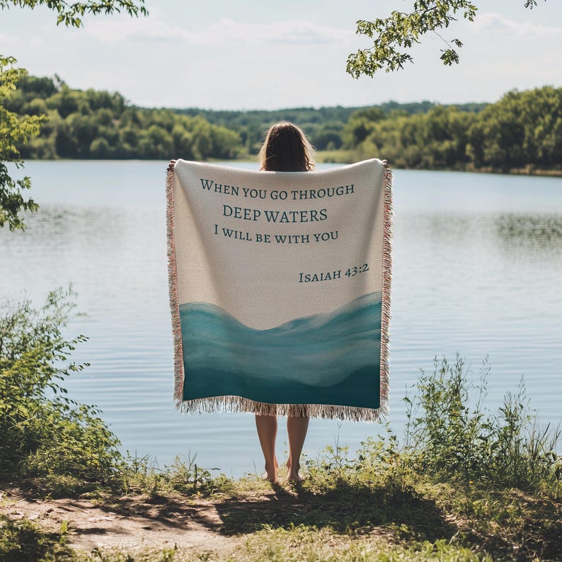 K&ouml;nnte beinhalten: Eine cremefarbene, gewebte Wohndecke mit einem blauen Aquarell-Wellenmuster am unteren Rand und dem in Dunkelblau gedruckten Text "WHEN YOU GO THROUGH DEEP WATERS I WILL BE WITH YOU ISAIAH 43:2", versehen mit Fransen an allen Seiten.
