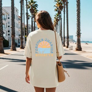 May include: Cream-colored t-shirt with a retro sunset graphic on the back. The design features a sun, waves, and the text "FOREVER CHASING SUNSETS" in pastel colors. The shirt is worn outdoors on a sunny day.