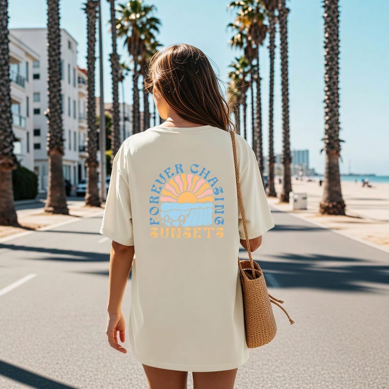 May include: Cream-colored t-shirt with a retro sunset graphic on the back. The design features a sun, waves, and the text "FOREVER CHASING SUNSETS" in pastel colors. The shirt is worn outdoors on a sunny day.