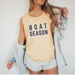 May include: A light yellow tank top with the words "BOAT SEASON" printed in navy blue. The tank top is sleeveless and has a rounded neckline. The person is wearing a light blue denim jacket and gray shorts.