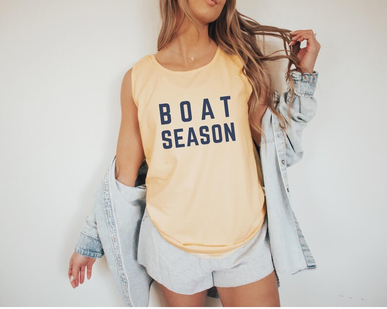 May include: A light yellow tank top with the words "BOAT SEASON" printed in navy blue. The tank top is sleeveless and has a rounded neckline. The person is wearing a light blue denim jacket and gray shorts.