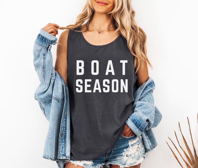 May include: Dark gray tank top with the words "BOAT SEASON" in white block letters. The tank top is worn with a light blue denim jacket and denim shorts. The image is taken in front of a white background.