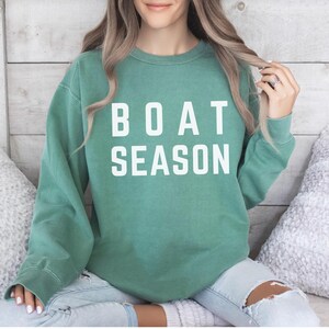 May include: A teal green sweatshirt with the words "BOAT SEASON" printed in white block letters. The long-sleeved crew neck top is worn with light wash jeans. The sweatshirt is a casual style.