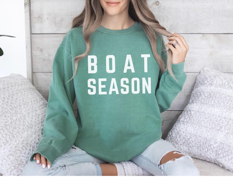 May include: A teal green sweatshirt with the words "BOAT SEASON" printed in white block letters. The long-sleeved crew neck top is worn with light wash jeans. The sweatshirt is a casual style.
