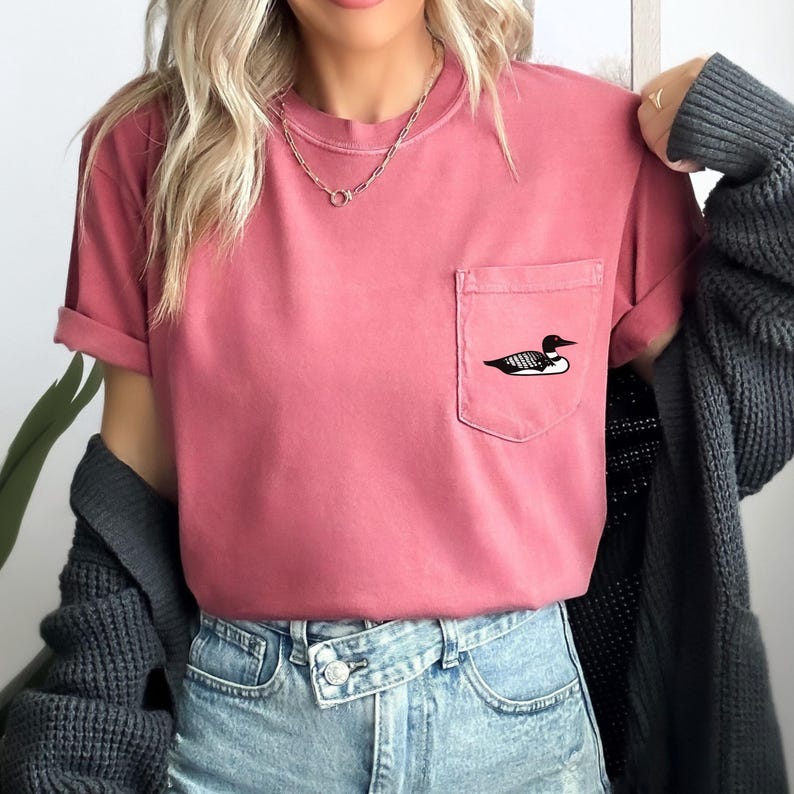 K&ouml;nnte beinhalten: Ein pinkfarbenes T-Shirt mit einer kleinen Tasche auf der linken Seite. Die Tasche hat ein schwarzes gesticktes Loon-Design.