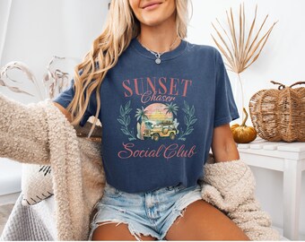 May include: A navy blue t-shirt with a vintage-inspired graphic design. The graphic features a sunset, palm trees, a jeep, and the text "Sunset Chaser Social Club".