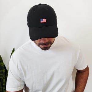Puede incluir: Gorra de b&eacute;isbol negra con una peque&ntilde;a bandera americana bordada en la parte delantera. La gorra la lleva una persona que viste una camiseta blanca. La gorra tiene una visera curva y una parte superior redondeada.