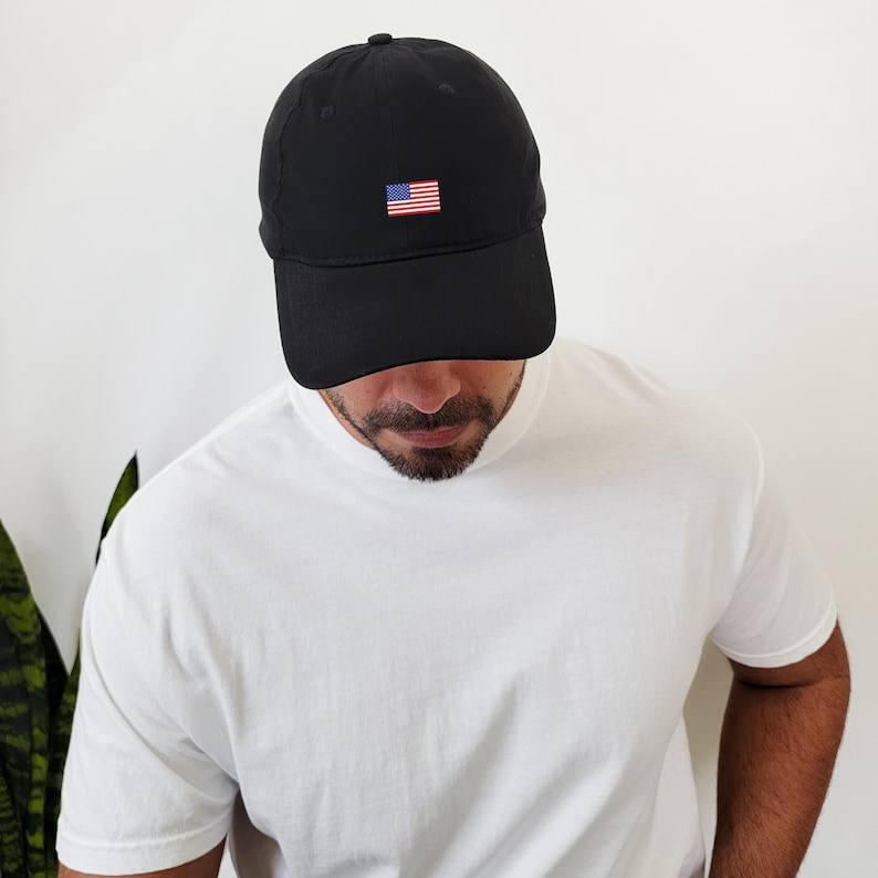 Puede incluir: Gorra de b&eacute;isbol negra con una peque&ntilde;a bandera americana bordada en la parte delantera. La gorra la lleva una persona que viste una camiseta blanca. La gorra tiene una visera curva y una parte superior redondeada.