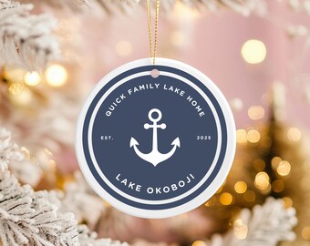 May include: A circular white ceramic ornament with a navy blue border and text. The text reads "Quick Family Lake Home" and "Lake Okoboji" with a white anchor in the centre. A gold string is attached for hanging.