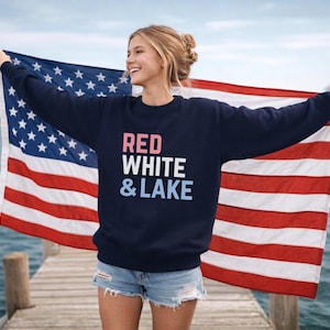 Könnte beinhalten: Marineblaues Sweatshirt mit dem Text "RED WHITE & LAKE" in Rot, Weiß und Blau. Die Person hält eine amerikanische Flagge mit roten und weißen Streifen und einem blauen Sternenfeld. Die Person steht auf einem Holzsteg.