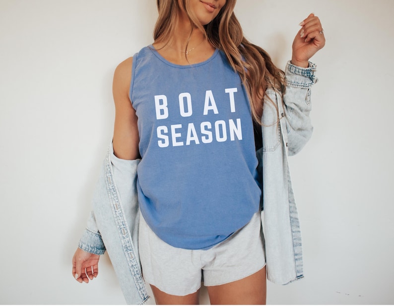 May include: A blue tank top with the words "BOAT SEASON" in white block letters. The tank top is paired with gray shorts and a light denim jacket. The outfit is casual and summery.