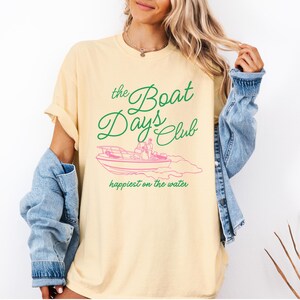 Pu&ograve; includere: T-shirt giallo chiaro con la scritta "the Boat Days Club" in verde sopra un'illustrazione rosa di una barca. La frase "happiest on the water" &egrave; sotto. Una giacca di jeans &egrave; indossata sulle spalle.