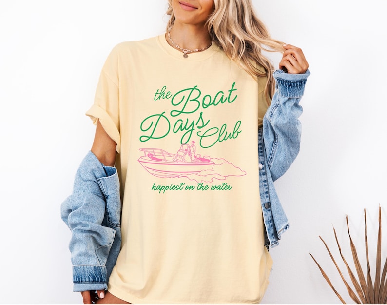 Pu&ograve; includere: T-shirt giallo chiaro con la scritta "the Boat Days Club" in verde sopra un'illustrazione rosa di una barca. La frase "happiest on the water" &egrave; sotto. Una giacca di jeans &egrave; indossata sulle spalle.