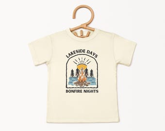Op de afbeelding: Een crèmekleurig T-shirt met een afbeelding van een kampvuur, bomen en een zon. De tekst "Lakeside Days" en "Bonfire Nights" staat op het shirt gedrukt.