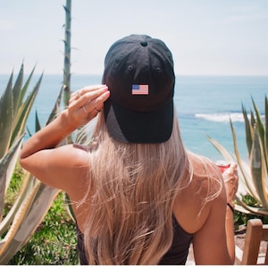 Puede incluir: Una gorra de b&eacute;isbol negra con un peque&ntilde;o parche de la bandera estadounidense es usada por una persona con cabello largo y rubio. La persona est&aacute; de espaldas a la c&aacute;mara, con vista al oc&eacute;ano y plantas verdes en el fondo.