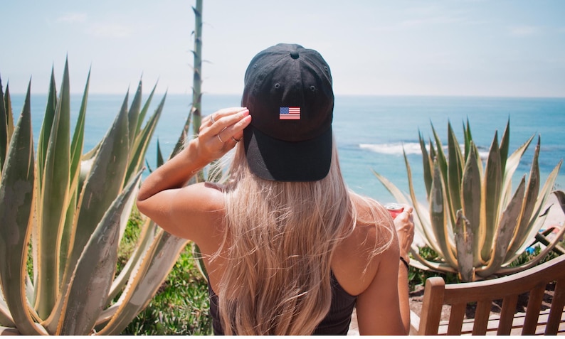 Puede incluir: Una gorra de b&eacute;isbol negra con un peque&ntilde;o parche de la bandera estadounidense es usada por una persona con cabello largo y rubio. La persona est&aacute; de espaldas a la c&aacute;mara, con vista al oc&eacute;ano y plantas verdes en el fondo.