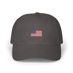 Puede incluir: Gorra de b&eacute;isbol gris oscuro con una peque&ntilde;a bandera estadounidense bordada en la parte delantera. La bandera presenta rayas rojas y blancas y un cuadrado azul con estrellas blancas.