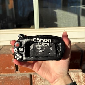 May include: A black, handmade camera-shaped sculpture with the word "Canon" in white. The camera's screen displays a black and white photo. The sculpture has red and white accents on the buttons and dials. The item is held in a hand.