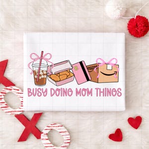 Puede incluir: Camiseta blanca con la frase rosa "Busy Doing Mom Things". El diseño incluye un café helado, una caja de nuggets de pollo, una tarjeta de crédito rosa y una caja de Amazon, todos adornados con lazos rosas.