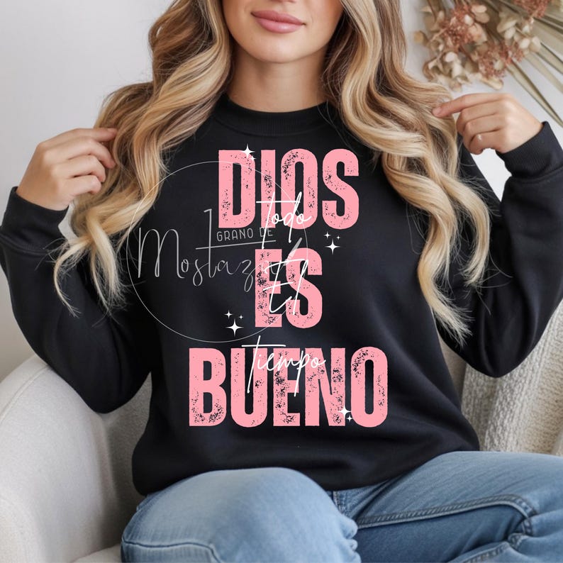 May include: Black crewneck sweatshirt with the text "DIOS ES BUENO" in pink. The sweatshirt also features the text "Mostaza" in a script font. The design includes small star accents.