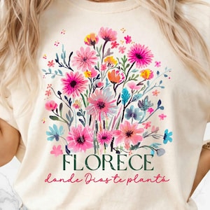 Könnte beinhalten: Cremefarbenes T-Shirt mit einem farbenfrohen Blumendesign in Rosa, Gelb, Orange und Blau. Das Design enthält den Text "FLORECE" über dem Satz "donde Dios te plantó" in einer Schreibschrift.