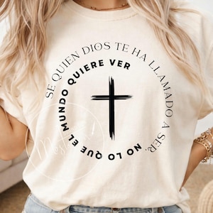 Puede incluir: Camiseta color crema con una cruz negra y el texto "SE QUIEN DIOS TE HA QUIERE VER, EL MUNDO NO LO QUE LLAMADO A SER." El diseño está centrado, con el texto en un patrón circular alrededor de la cruz.