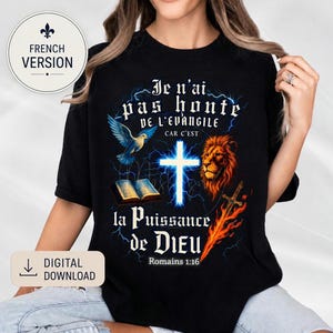 Puede incluir: Camiseta negra con un diseño gráfico que presenta una cruz, un león, una paloma y un libro abierto. El texto dice "Je n'ai pas honte de l'Evangile" y "la Puissance de DIEU" en blanco. Un logo de "French Version" está en la parte superior izquierda.