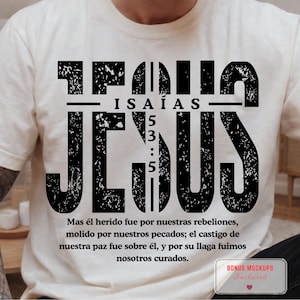 May include: A cream-coloured t-shirt featuring a large, distressed black graphic that reads JESUS in bold letters. Centered within the text is the reference ISAÍAS 5:3:5. Below, the Spanish text reads: Mas él herido fue por nuestras rebeliones.
