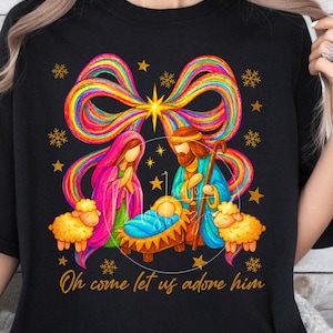 Puede incluir: Camiseta negra con una colorida ilustración del Nacimiento. La escena muestra a María, José y el niño Jesús, coronada por un arcoíris y una estrella. El texto dice "Oh come let us adore him."