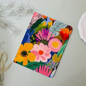 May include: A colorful notebook cover with a floral design. The cover features a variety of flowers in shades of pink, yellow, red, and purple, as well as green leaves. The background is a dark blue.