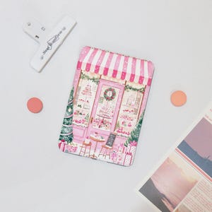 Op de afbeelding: Een roze en witte decoratieve blikken doos met een bakkerijscène met taarten, cadeaus en een krans. Bovenop een witte clip met de tekst "Near Uniform". Twee kleine, ronde, perzik-kleurige objecten aan de linker- en rechterkant.